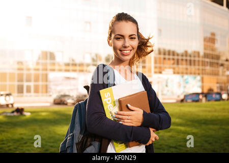 Portrait d'une joyeuse fille étudiante brune debout avec sac à dos et des livres et à la recherche à l'extérieur de l'appareil photo Banque D'Images