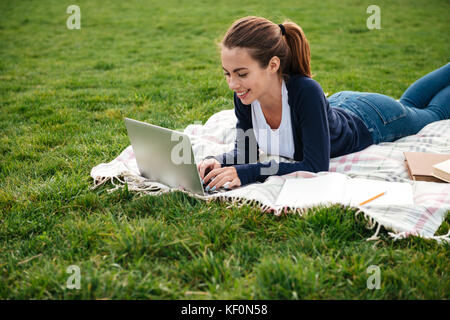 Portrait of a smiling beautiful student girl faire leurs devoirs sur ordinateur portable tout en étant allongé sur l'herbe au parc Banque D'Images