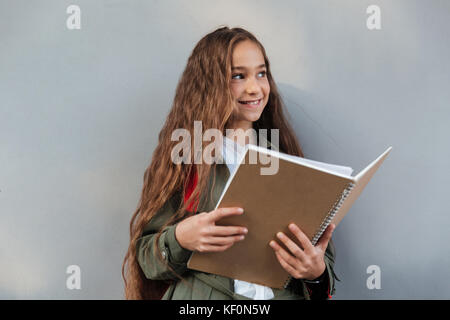 Happy brunette lycéenne aux cheveux longs habillés en vêtements chauds et à l'écart de l'ordinateur portable holding sur fond gris Banque D'Images