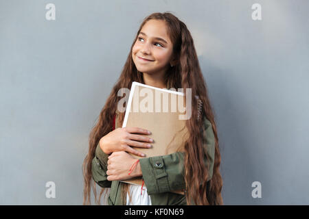 Smiling brunette lycéenne aux cheveux longs habillés en vêtements chauds et à l'écart de l'ordinateur portable holding sur fond gris Banque D'Images