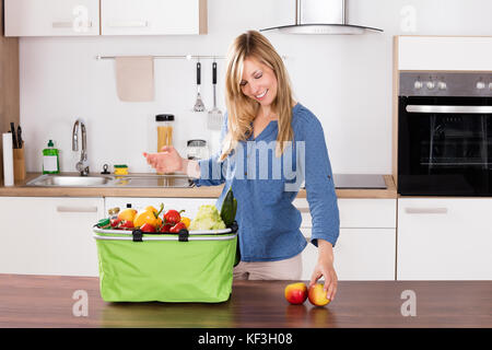 Souriante jeune femme dépose les pommes de sac d'épicerie dans la cuisine Banque D'Images