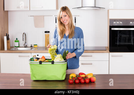 Souriante jeune femme dépose des raisins de sac d'épicerie dans la cuisine Banque D'Images