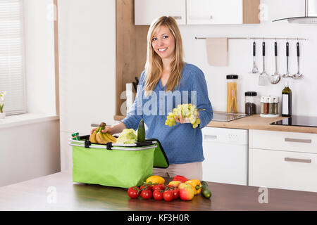 Souriante jeune femme dépose des raisins de sac d'épicerie dans la cuisine Banque D'Images