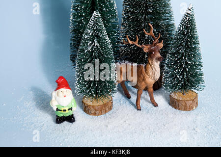 Mise en scène de sapins artificiels complet comme une petite forêt enneigée arbre avec une figurine à l'intérieur de rennes et d'un nain de jardin à côté sur un fond bleu. Banque D'Images