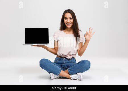 Portrait of a happy girl holding blank écran ordinateur portable et montrant ok assis geste isolé sur fond blanc Banque D'Images