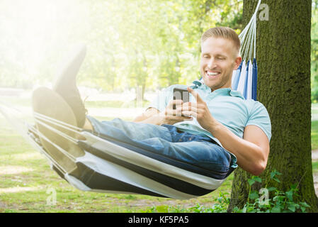 Young happy man using mobile phone while relaxing in hammock at garden