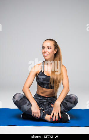 Sportive et mettre en place la beauté. belle femme sportswear looking at camera avec smile while sitting on exercise mat contre l'arrière-plan gris Banque D'Images