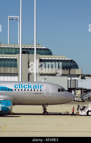 Avant du fuselage d'un Airbus A320-200 clickair, un remorqueur avec un attelage, un travailleur de l'aéroport orange portant un tabard et ear-humains avec une passerelle et te Banque D'Images