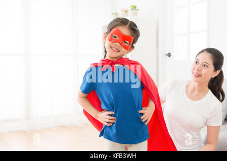 Belle petite jeunesse kid girl face à l'appareil photo porte manteau rouge avec masque jouer en tant que super-héros et de beauté smiling mother la regarder. Banque D'Images