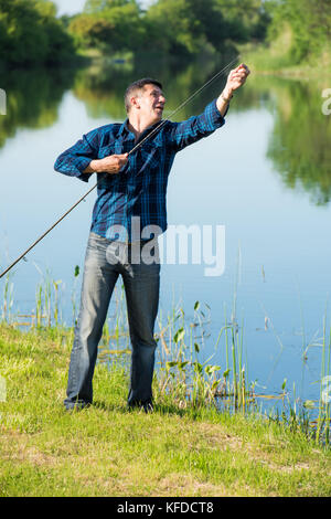 Personnes âgées, mais l'homme est toujours actif fixant sa verge tout en se préparant à aller à la pêche. L'homme est debout sur l'herbe verte de la rivière. Une forêt non focalisé Banque D'Images