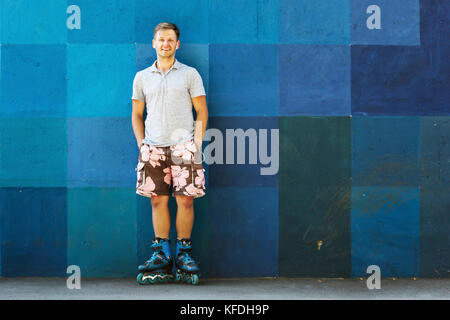 Portrait of happy handsome young en patins à rouleaux debout contre un mur bleu. Banque D'Images