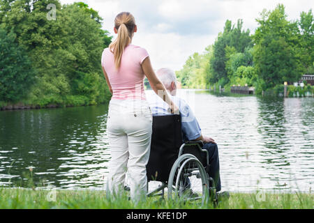 Vue arrière d'une jeune femme avec son père handicapé sur fauteuil roulant à au lac Banque D'Images