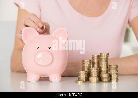 Close-up of Woman Inserting Coin In Piggybank en face de pièces empilées Banque D'Images