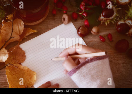 La main féminine est titulaire d'un crayon sur une feuille de papier blanc, tasse de thé de thé de rose musquée et bouquet de rose hips et châtaignes,feuilles d'automne sur l'woode Banque D'Images