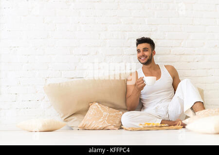 Jeune homme hispanique s'asseoir sur des coussins marbre, happy smiling guy avec plateau de petit-déjeuner chambre espace copie rechercher Banque D'Images