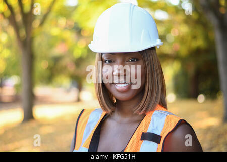 Une femme noire construction worker wearing a white hat et gilet orange qui travaillent à l'extérieur un parc de la ville Banque D'Images