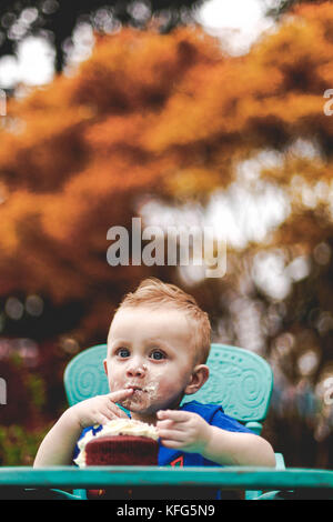 Wide eyed baby boy eating cake dans sa chaise haute. bleu arbres en arrière-plan flou. Banque D'Images