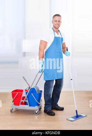 Nettoyage Entretien du parquet avec une vadrouille et un panier avec deux seaux pour l'eau et de désinfectant s'arrêtant de sourire à la caméra en tant qu'il se passe à propos de h Banque D'Images