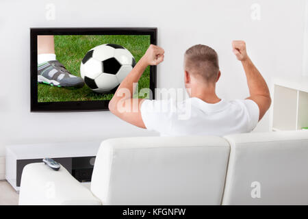 Heureux fan de foot un match à la télévision tenant un ballon au-dessus de sa tête comme il est assis sur un canapé confortable dans son salon Banque D'Images