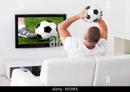 Heureux fan de foot un match à la télévision tenant un ballon au-dessus de sa tête comme il est assis sur un canapé confortable dans son salon Banque D'Images