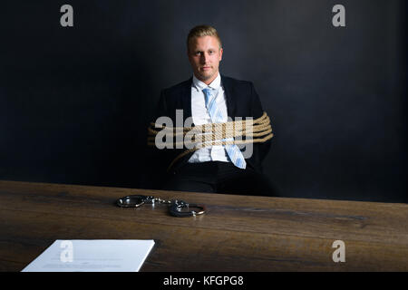 Portrait de jeune homme d'affaires attaché avec corde assis devant la table avec menottes placées dessus Banque D'Images