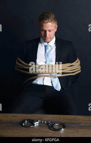 Portrait de jeune homme d'affaires attaché avec corde assis devant la table avec menottes placées dessus Banque D'Images