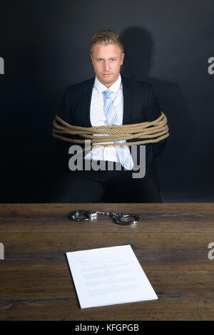 Portrait de jeune homme d'affaires attaché avec corde assis devant la table avec menottes placées dessus Banque D'Images