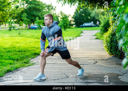 Un athlète masculin se réchauffe avant de faire du jogging dans le parc. Écoute de la musique sur le casque. Banque D'Images