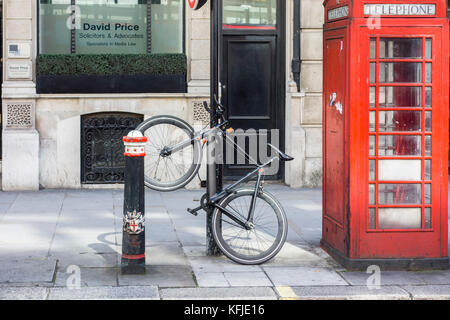 En stationnement vélo sécurisé verrouillé à un bollard post à côté d'un téléphone rouge fort, Ville de London, UK Banque D'Images