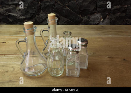 L'huile d'olive, vinaigre de bouteilles en verre, salières et poivrières mis sur table en bois, contre un mur en pierre noire Banque D'Images