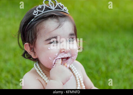 Smiling happy baby girl portrait premier anniversaire anniversaire partie. lécher les doigts avec du rose sale face gâteau. princess tiara costume Banque D'Images