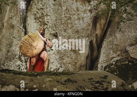 Strong warrior dans gladiator armor couvrant avec la protection en flèches et posant avec épée en étant debout sur les rochers. Banque D'Images