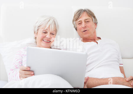 Portrait Of Happy Senior Couple Lying On Bed Looking At Laptop Banque D'Images