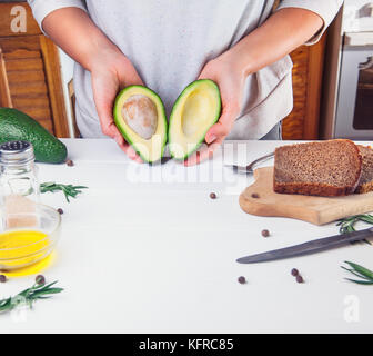 Femme tenant les moitiés d'avocat frais sous une table en bois blanc avec des ingrédients pour faire des sandwichs. Le mode de vie sain, l'alimentation, les graisses végétales concept backg Banque D'Images
