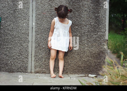 Triste petit enfant (fille) debout contre un mur, regardant vers le bas. Humeur malheureuse, tir à l'extérieur Banque D'Images