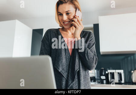 Femme parlant au téléphone mobile et son ordinateur portable d'exploitation alors qu'à la maison. Femme d'affaires qui fréquentent au travail de bureau de sa cuisine. Banque D'Images