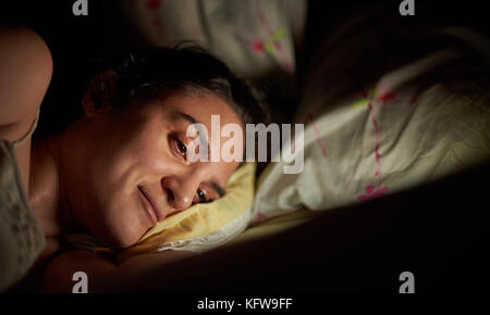 Smiling woman using tablet au lit avant de dormir. La lumière vive à partir de la tablette numérique sur fille face au lit Banque D'Images