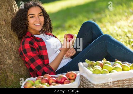 Beautiful happy mixed race african american girl adolescent femme jeune femme dans un verger biologique en mangeant une pomme rouge, souriant avec dents parfait lea Banque D'Images