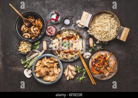 La cuisson des aliments asiatiques. wok avec sauté de poulet nouilles et légumes ingrédients avec des épices, sauces et baguettes sur fond rustique foncé Banque D'Images