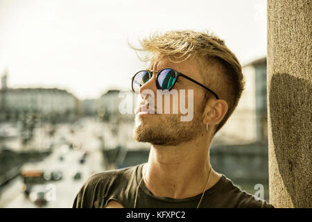 Blonde aux yeux bleus, beau jeune homme appuyé contre un mur en pierre et en plein air à la recherche d'un côté à Turin, Italie Banque D'Images