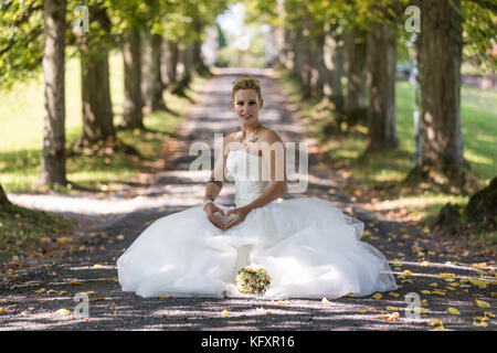 Jeune femme blonde en robe de mariée blanche, assis et avec coeur geste, Suisse Banque D'Images