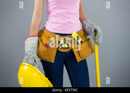 Section intermédiaire de femme avec ceinture d'outils et holding hard hat contre l'arrière-plan gris Banque D'Images