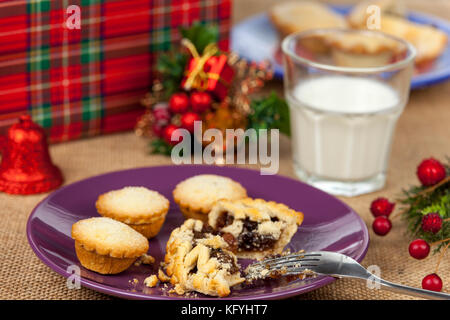 Broken mince pie sur une plaque pourpre avec un verre de lait sur une table de Noël avec une nappe de Hesse Banque D'Images