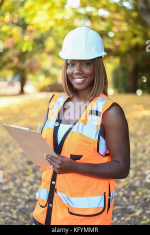 Une femme noire construction worker wearing a white hat et gilet orange qui travaillent à l'extérieur un parc de la ville Banque D'Images