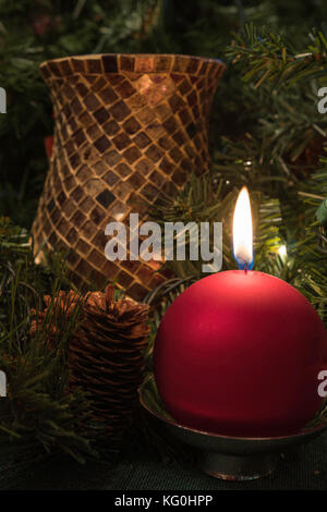 Affichage de Noël avec une bougie boule rouge entouré de verdure, des pommes de pin et un autre porte-bougie à l'arrière-plan Banque D'Images