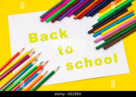 L'inscription - retour à l'école, sur une feuille de papier blanc entouré par des crayons, marqueurs, peintures sur un fond jaune lumineux Banque D'Images