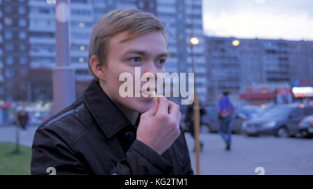 Guy mange des pommes de terre frites dans la rue, sur le banc. Homme mangeant des frites, pommes de terre frites, chips, fast food malsain. jeune homme mangeant un hamburger avec des pommes de terre frites dans un café de nourriture de rue. Restauration rapide Banque D'Images