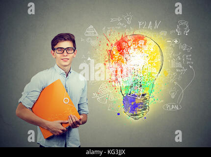 Portrait of young happy smiling man with orange ordinateur portable dossier près d'un mur de béton avec une ampoule lumineuse idée d'entreprise sketch isolé sur ba gris Banque D'Images