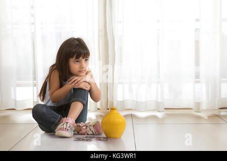 Petite fille assise sur le plancher avec de l'argile tirelire et pièces Banque D'Images