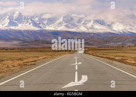 Belle vue de l'automne avec une route asphaltée à travers la steppe de la montagne couverte de neige, les arbres d'or sur la route et fluffy clouds Banque D'Images